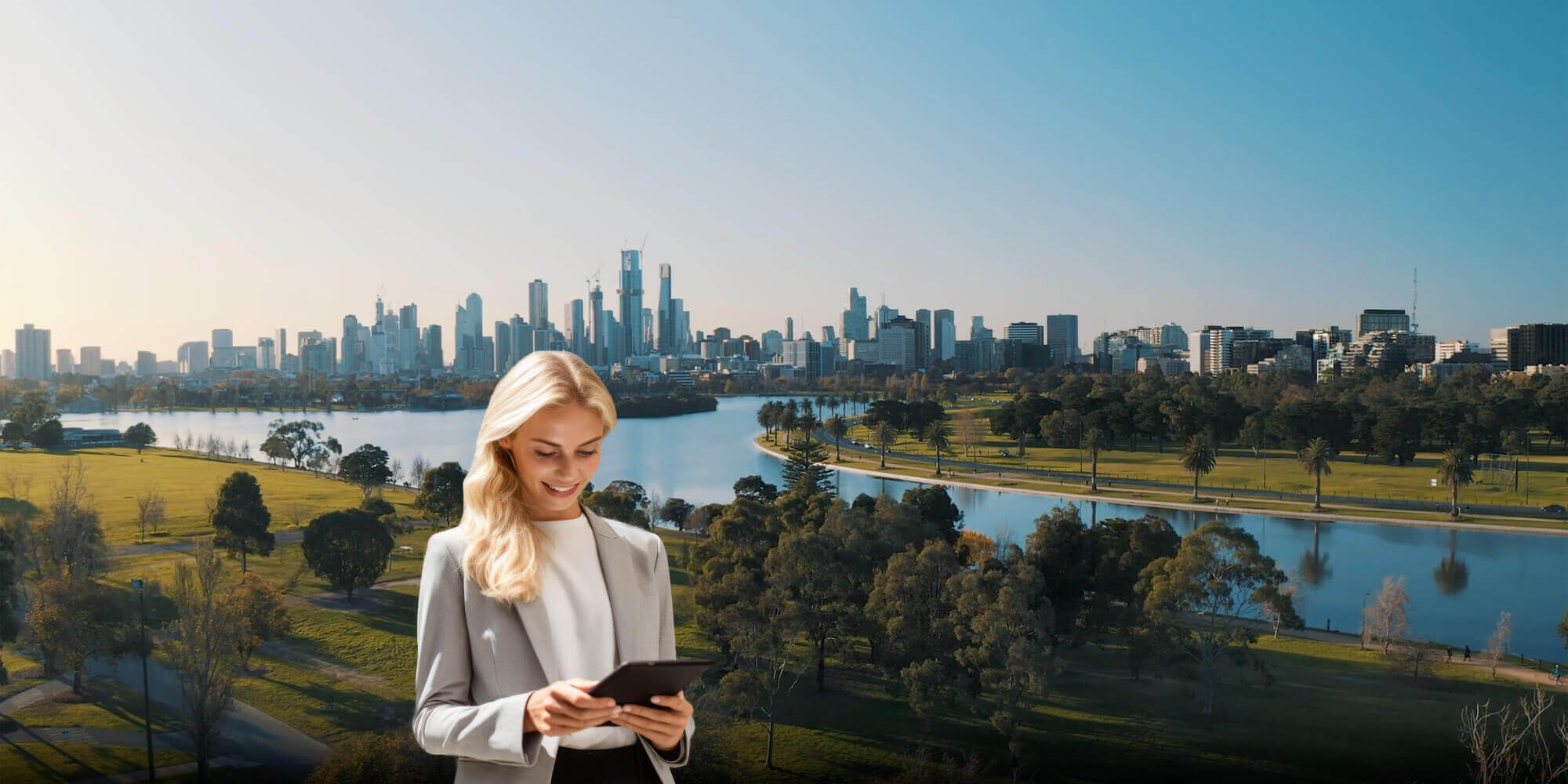 Eine Business - Frau im grauen Hosenanzug und weißen T- Shirt steht mit einem Tablet und arbeitet. Im Hintergrund ist eine größere Stadt mit Fluss, Straße, Grünflächen und Bäumen zu sehen.