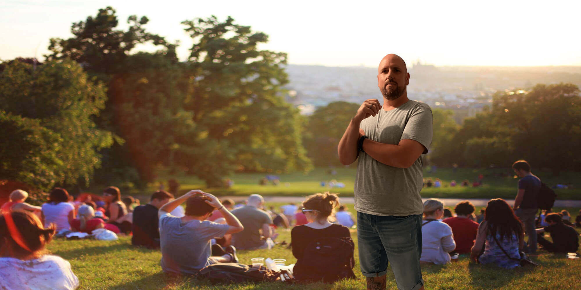 Mann in Freizeitkleidung blickt auf eine Wiese mit Menschen im Hintergrund, die teilweise sitzen und stehen.