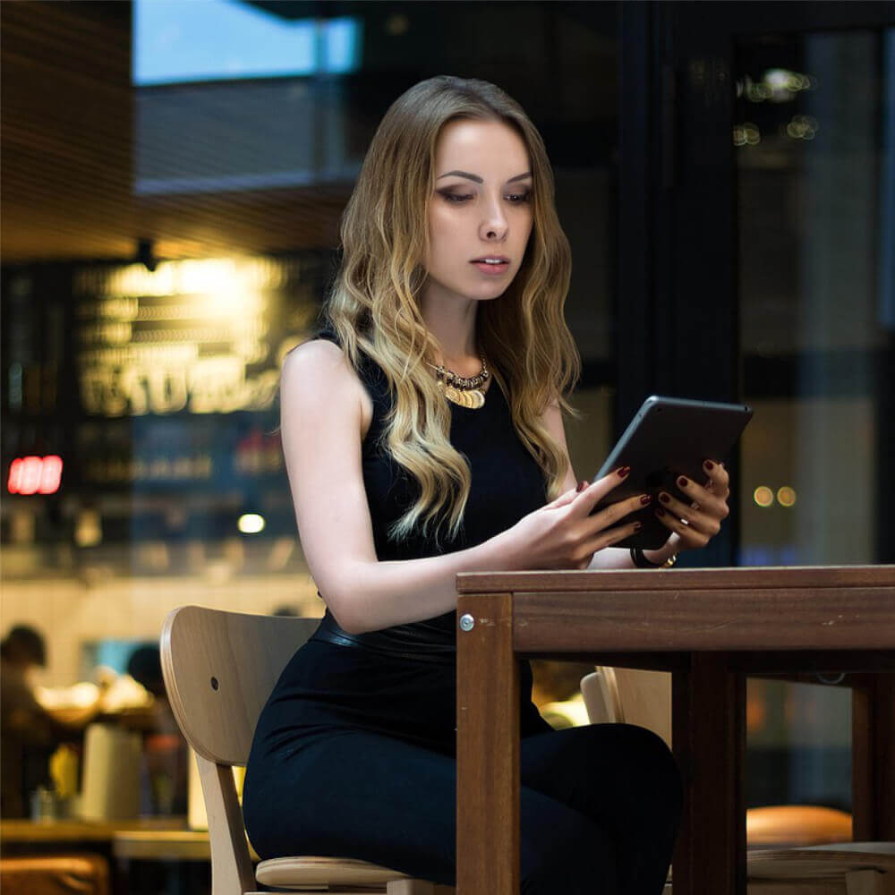 Eine elegant gekleidete Frau in Schwarz sitzt auf einem Hocker und konzentriert sich auf ein Tablet, während sie in einem stilvollen Lokal sitzt.