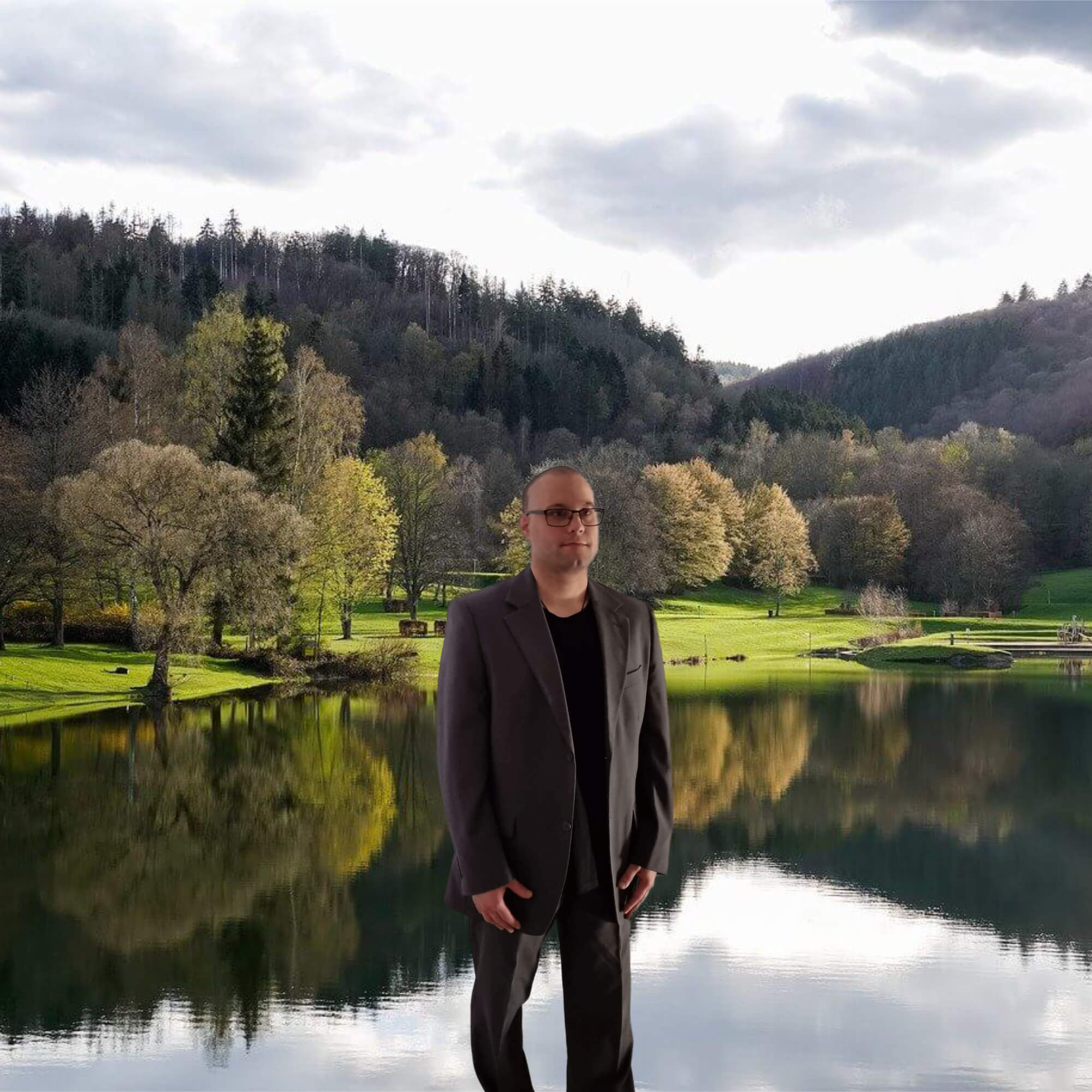 Ein Mann - DDr. Mario-Franz Wilfinger, MBA MA MSc in einem grauen Anzug und einem schwarzen T-Shirt steht vor einer Naturlandschaft mit grüner Wiese, Bäumen und einem Natursee.