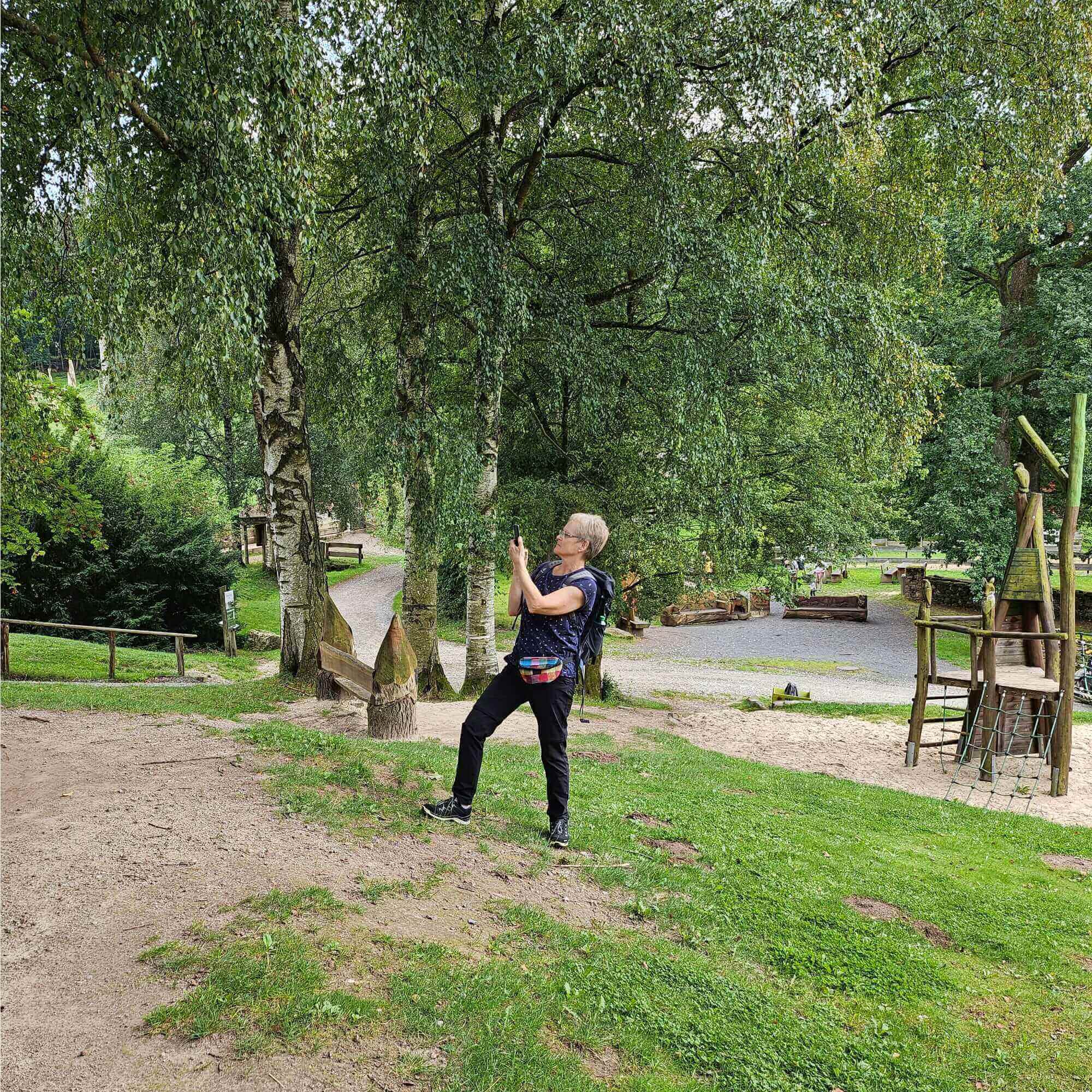 Eine Frau - Helga Monika Matheus mit Brille und kurzen Haaren in Sportbekleidung, die in einem Freizeit Park ein Foto mit ihrem Smartphone macht.