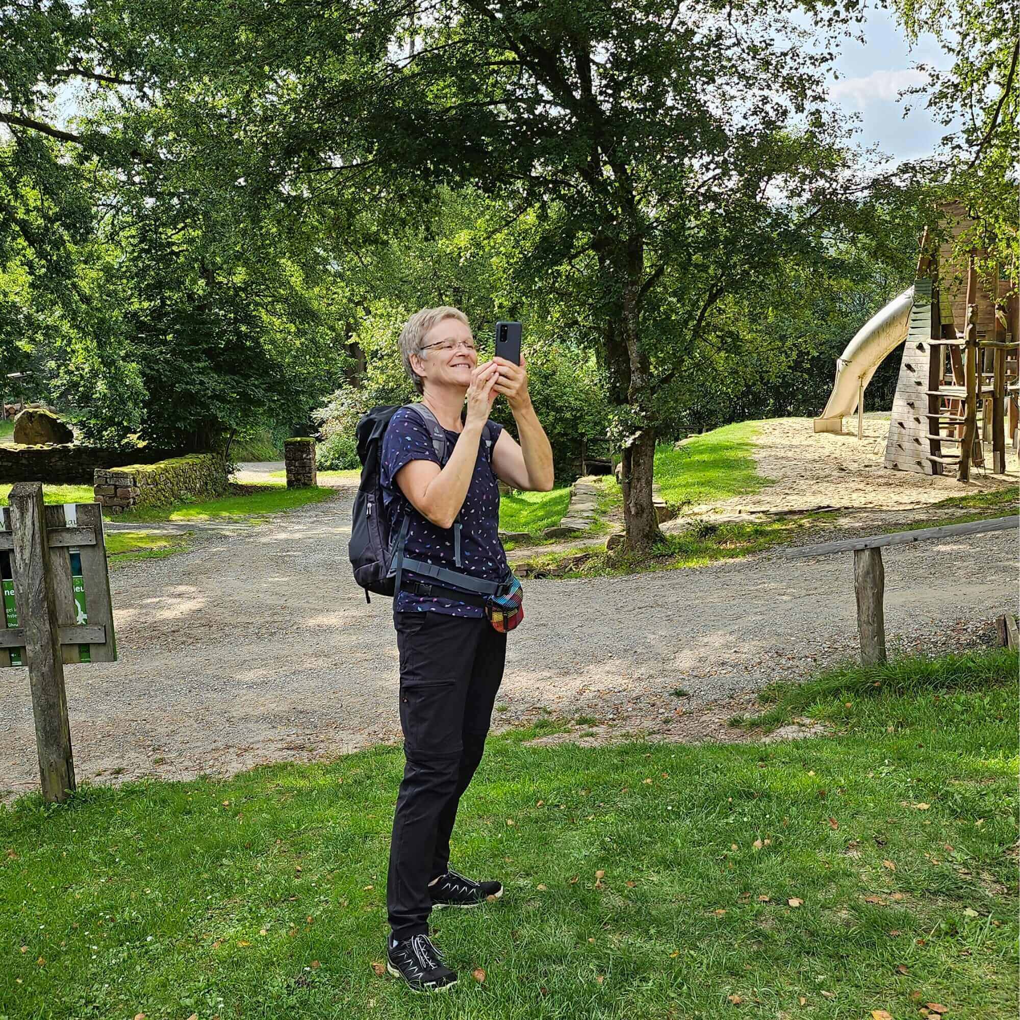 Eine Frau - Helga Monika Matheus mit Brille und kurzen Haaren in Sportbekleidung, die in einem Park ein Foto mit ihrem Smartphone macht.