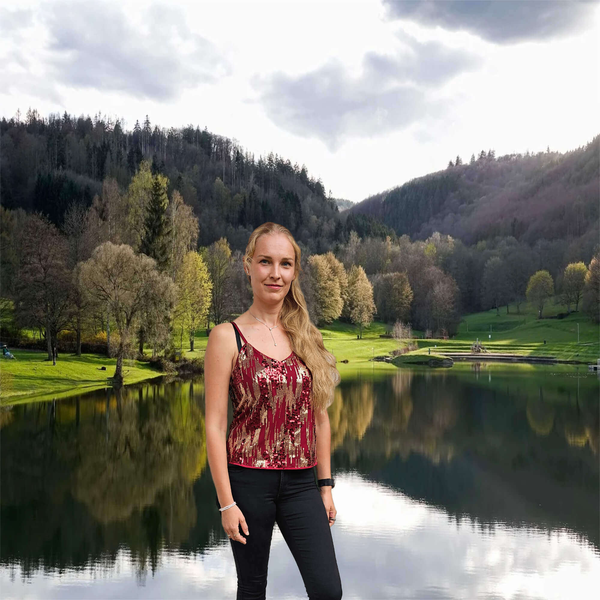 Eine Frau - Inger Maren Matheus, B.Ed M.Ed in schwarzer Hose und rotem festlichen Top steht vor einer Naturlandschaft mit grüner Wiese, Bäumen und einem Natur See.