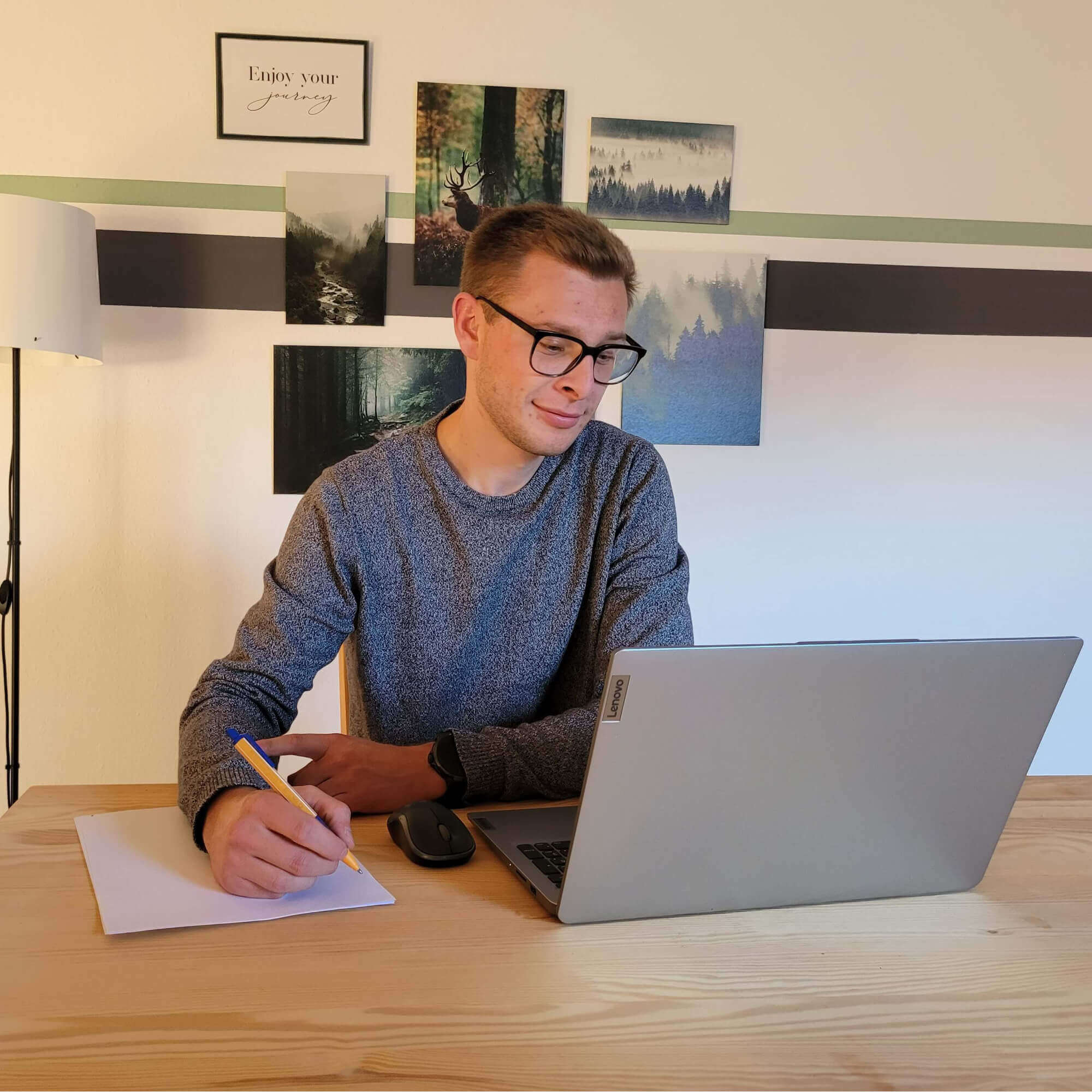 Ein Mann - Aron Matheus mit Brille arbeitet konzentriert im Home - Office mit einem Laptop, dass Smartphone und der Notizblock liegen auf dem Tisch und er hält einen Stift.