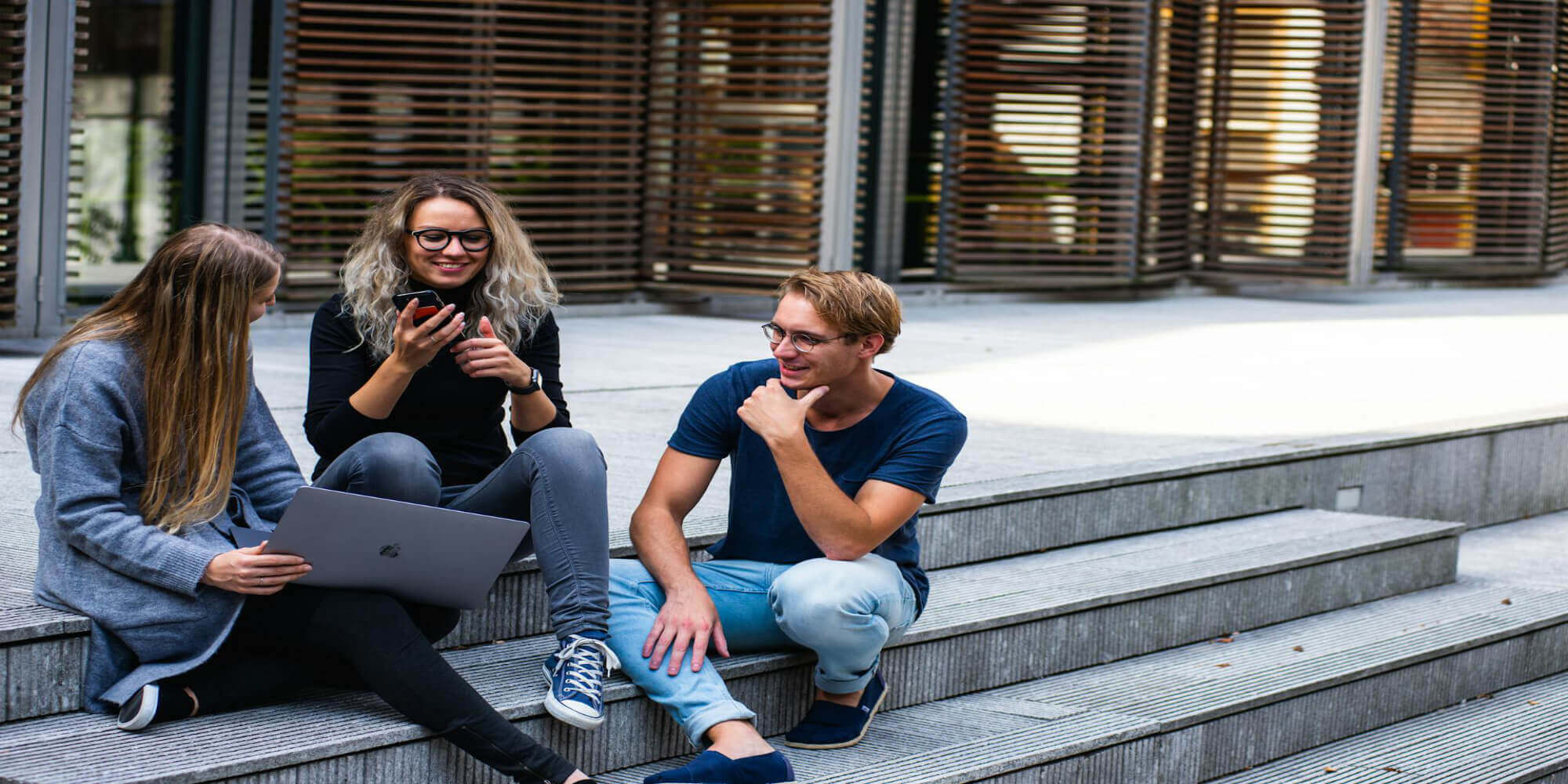 Drei Personen, sportlich gekleidet, zwei Frauen mit langen blonden und braunen Haaren und ein Mann mit kurzen braunen Haaren, sitzen auf einer modernen Stiege. Eine Frau hält einen Laptop, die andere Frau hält ein Smartphone, während diese sich untereinander unterhalten. Im Hintergrund ist ein modernes Gebäude zu sehen.