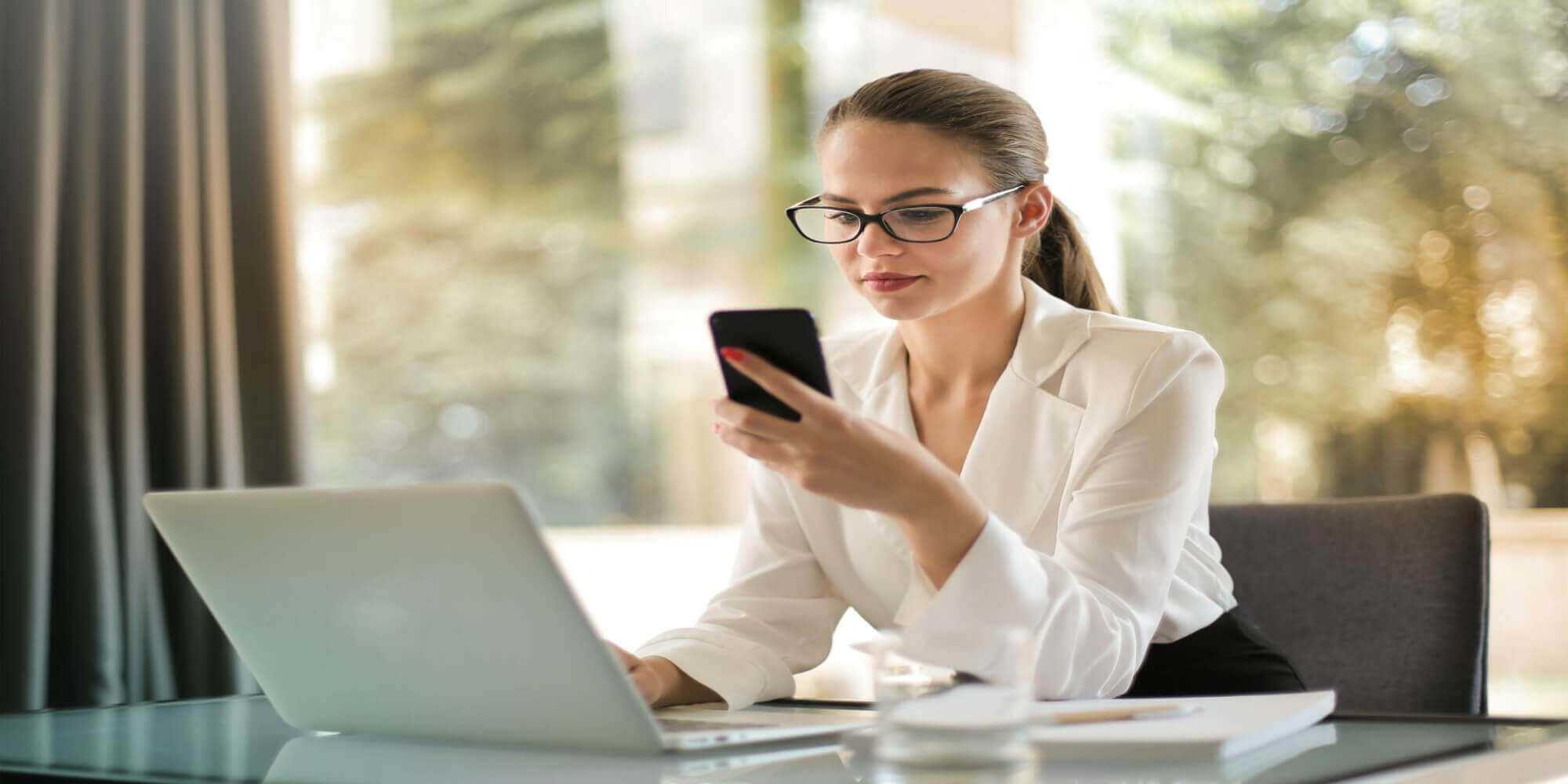 Eine Frau mit blonden und braunen langen Haaren, die eine Brille und einen weißen Business - Hosenanzug trägt, sitzt auf einem Sessel in einem modernen Raum. Vor ihr auf einem Tisch steht ein Laptop und sie hält ein Smartphone in den Händen, auf das sie konzentriert blickt.