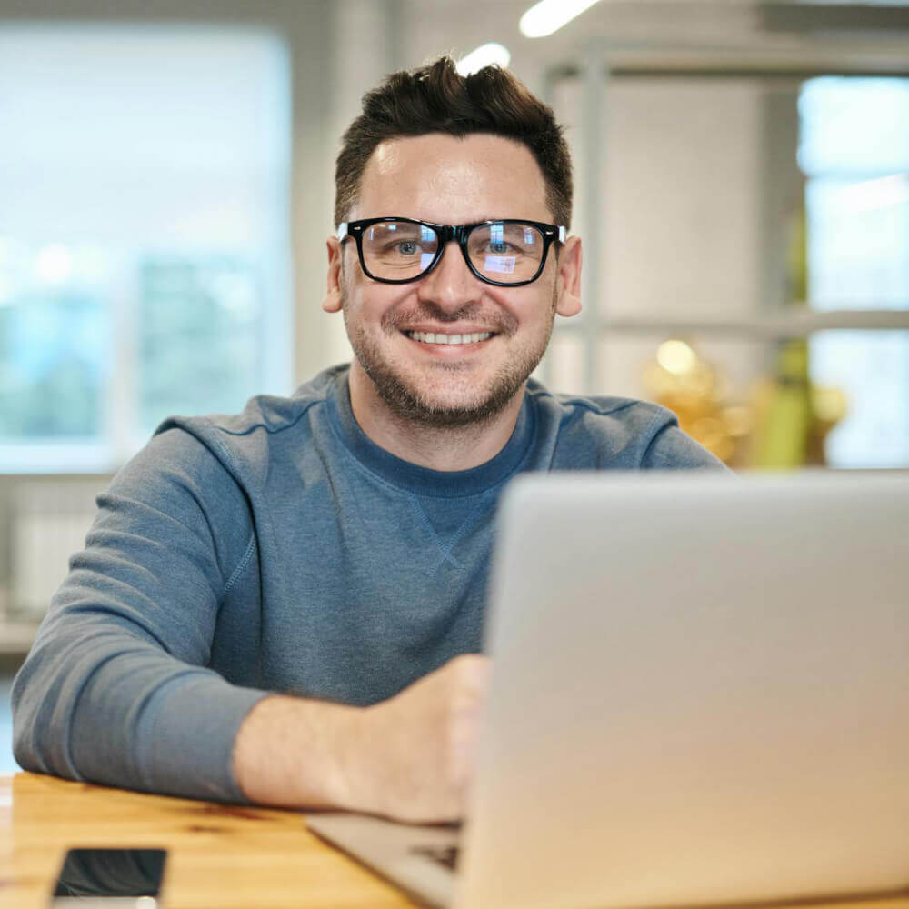 Ein Mann sitzt in einem modernen Bürogebäude auf einem Sessel, arbeitet an einem Laptop und verwendet gleichzeitig ein Smartphone.