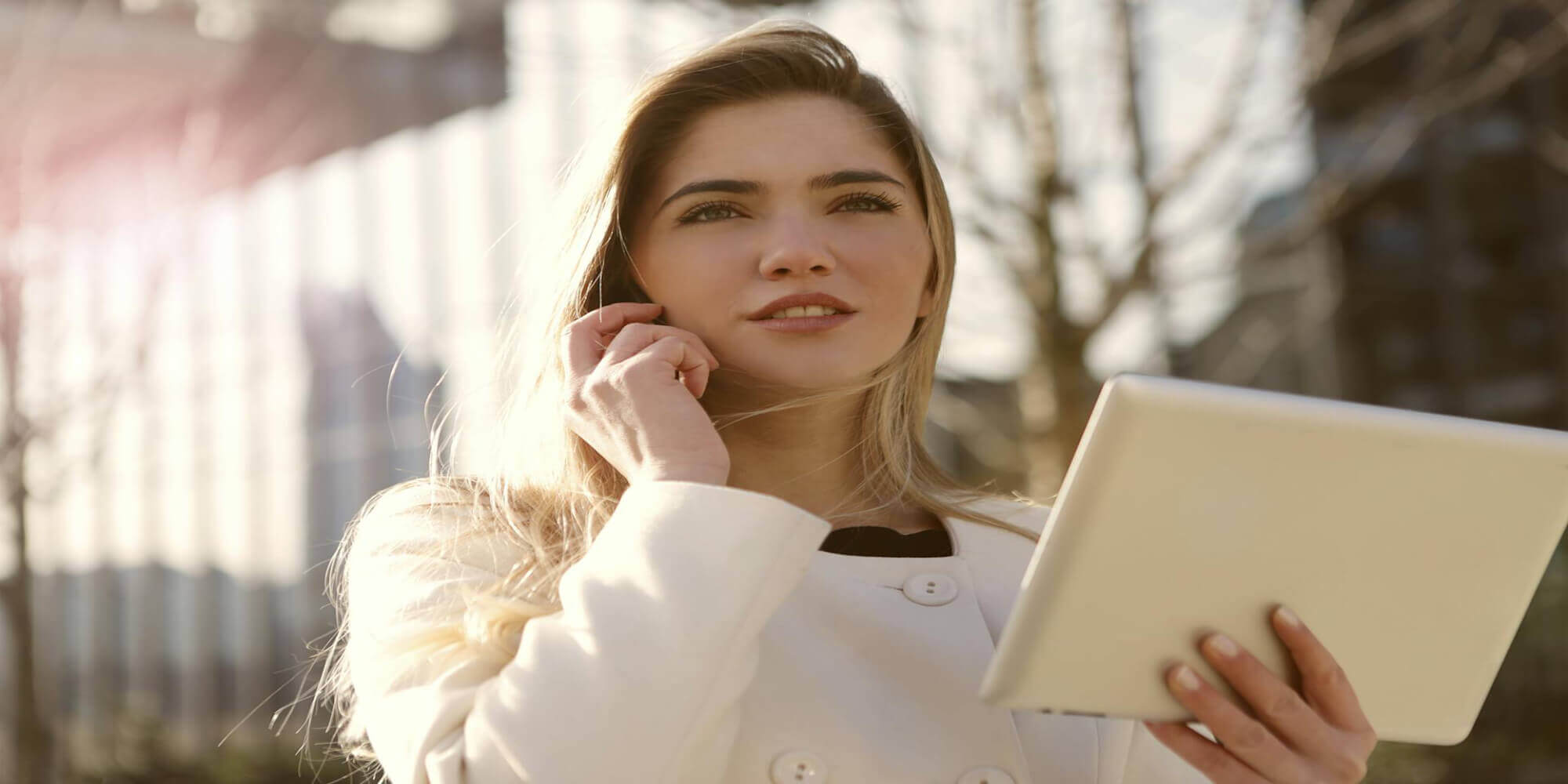 Eine hübsche Frau mit langen blond - braunen Haaren steht auf einem Platz, hält ein Smartphone in der einen Hand und telefoniert, während sie in der anderen Hand ein Tablet hält. Sie lächelt leicht und schaut fokussiert in die Ferne.