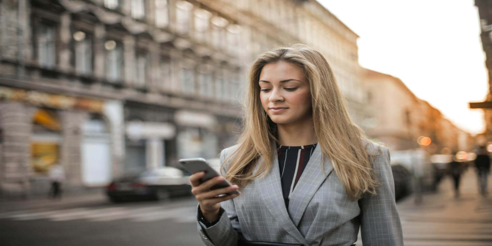 Eine Frau mit langen blonden Haaren trägt einen grauen Blazer und ein elegantes T - Shirt, während sie auf dem Gehsteig steht und auf ihr Smartphone schaut.