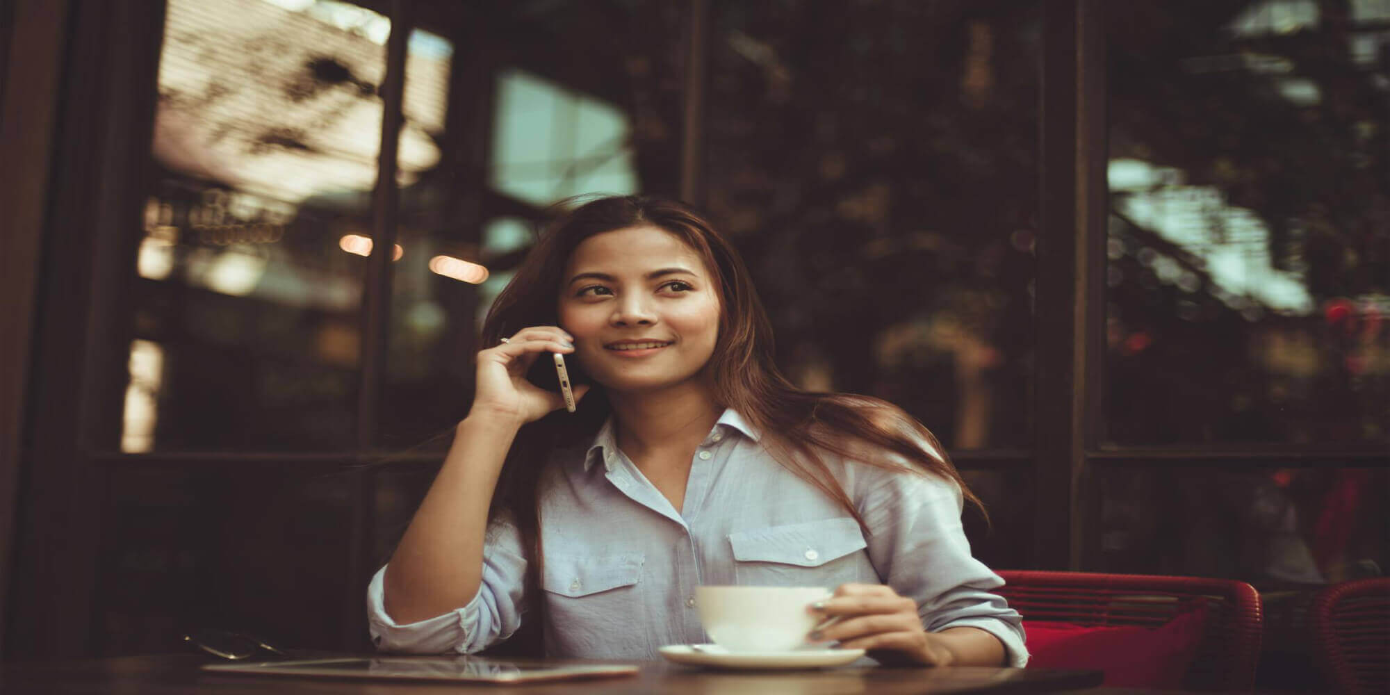 Eine hübsche Frau mit langen braunen Haaren sitzt in einem modernen und eleganten Restaurant, trinkt einen Kaffee und telefoniert mit einem Smartphone.