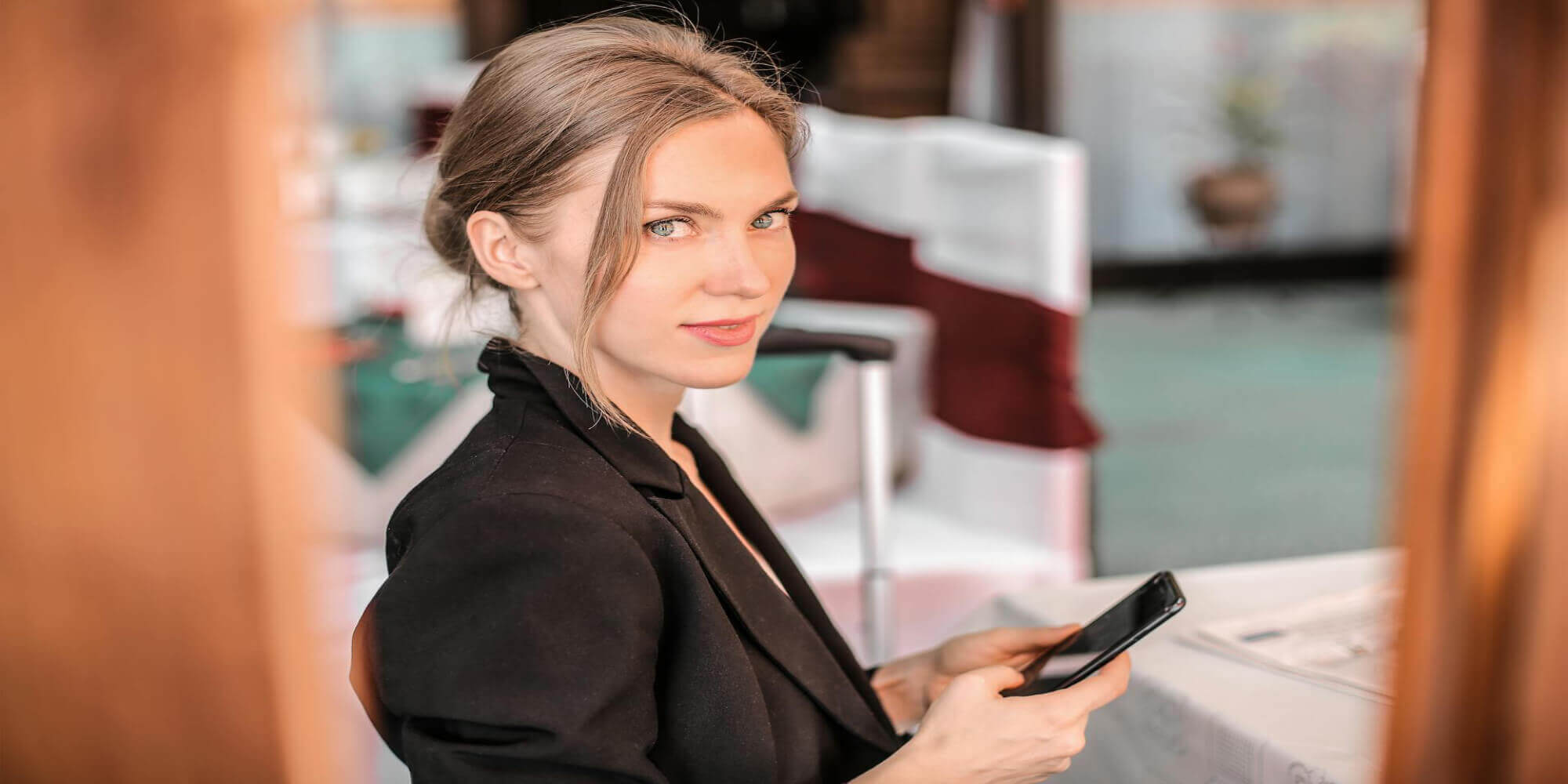 Eine hübsche Frau mit aufgesteckten braun - blonden Haaren trägt einen schwarzen Hosenanzug und hält ein Smartphone in den Händen. Sie steht in einem Raum und schaut in die Ferne.