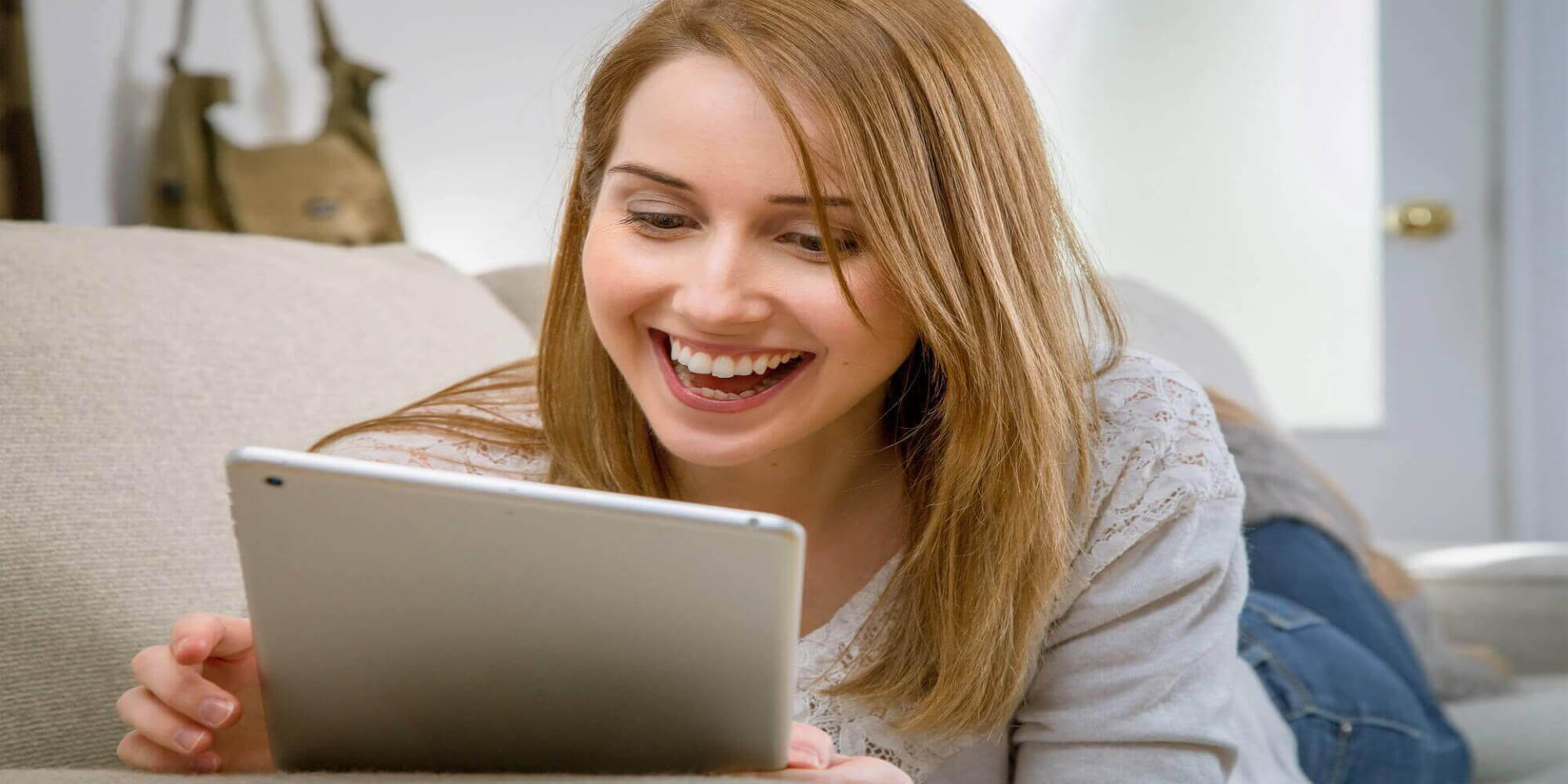 Eine Frau mit blonden Haaren in einem modernen, hellen Raum auf einer Couch sitzt und mit einem Tablet arbeitet.