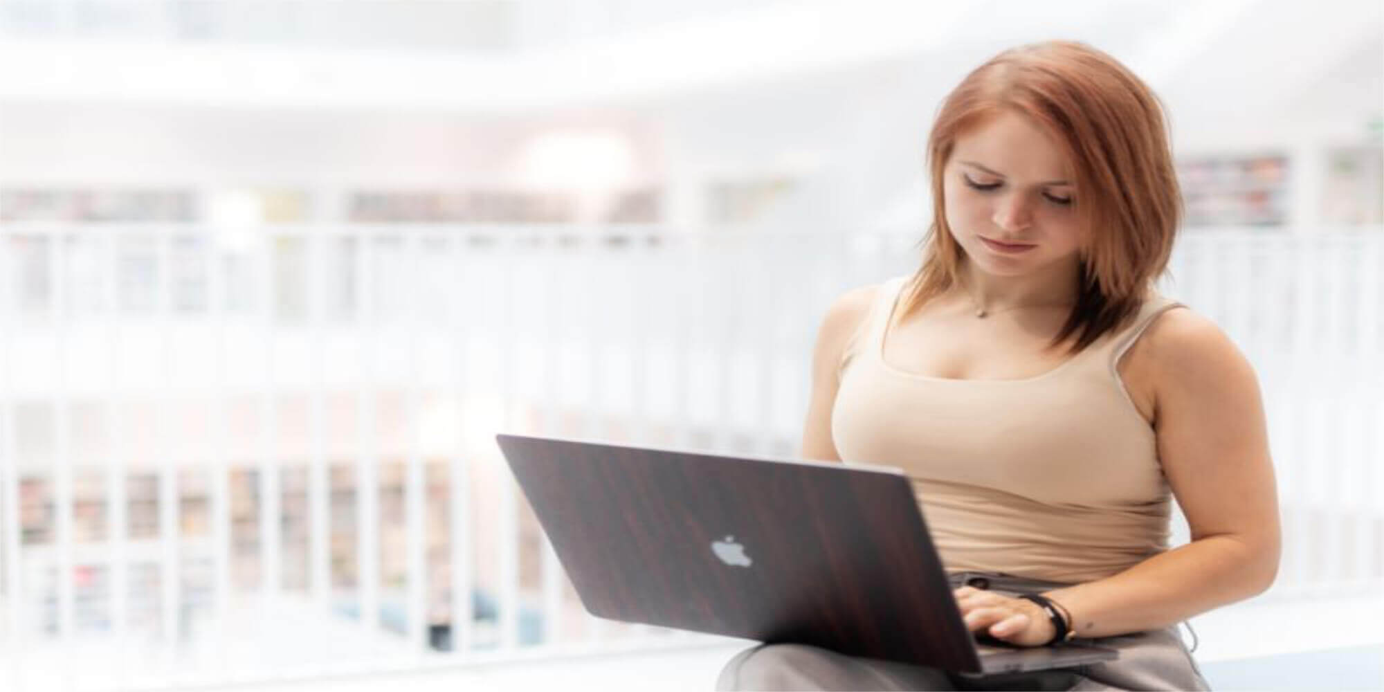 Eine Frau - Judith Langer mit langen roten Haaren in einem modernen Business- Look sitzt auf einem Sessel in einem zeitgemäßen Gebäude und arbeitet mit einem Laptop. Sie trägt einen Smart Ring.