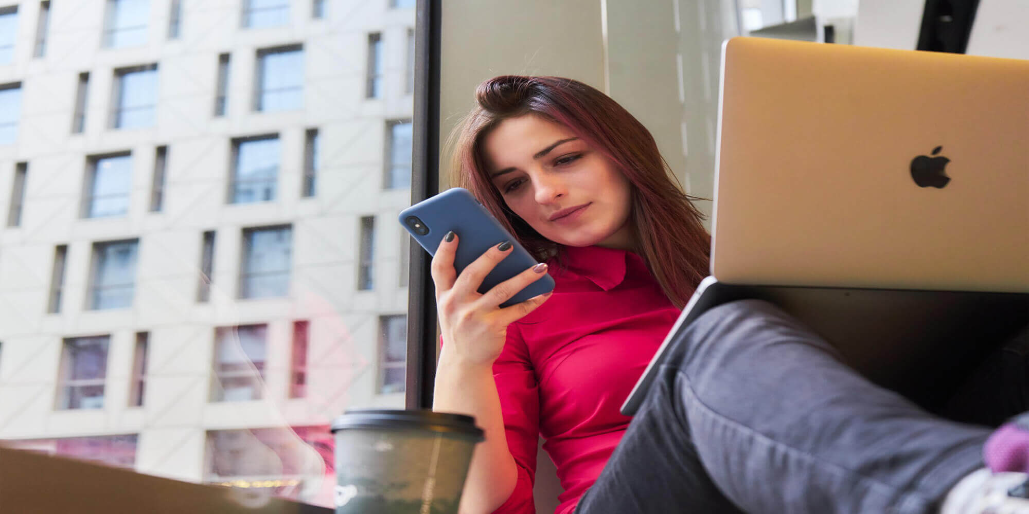 Frau mit rot-schwarzen langen Haaren sitzt am Fenster und arbeitet mit Laptop, während sie auf ihr Smartphone schaut.