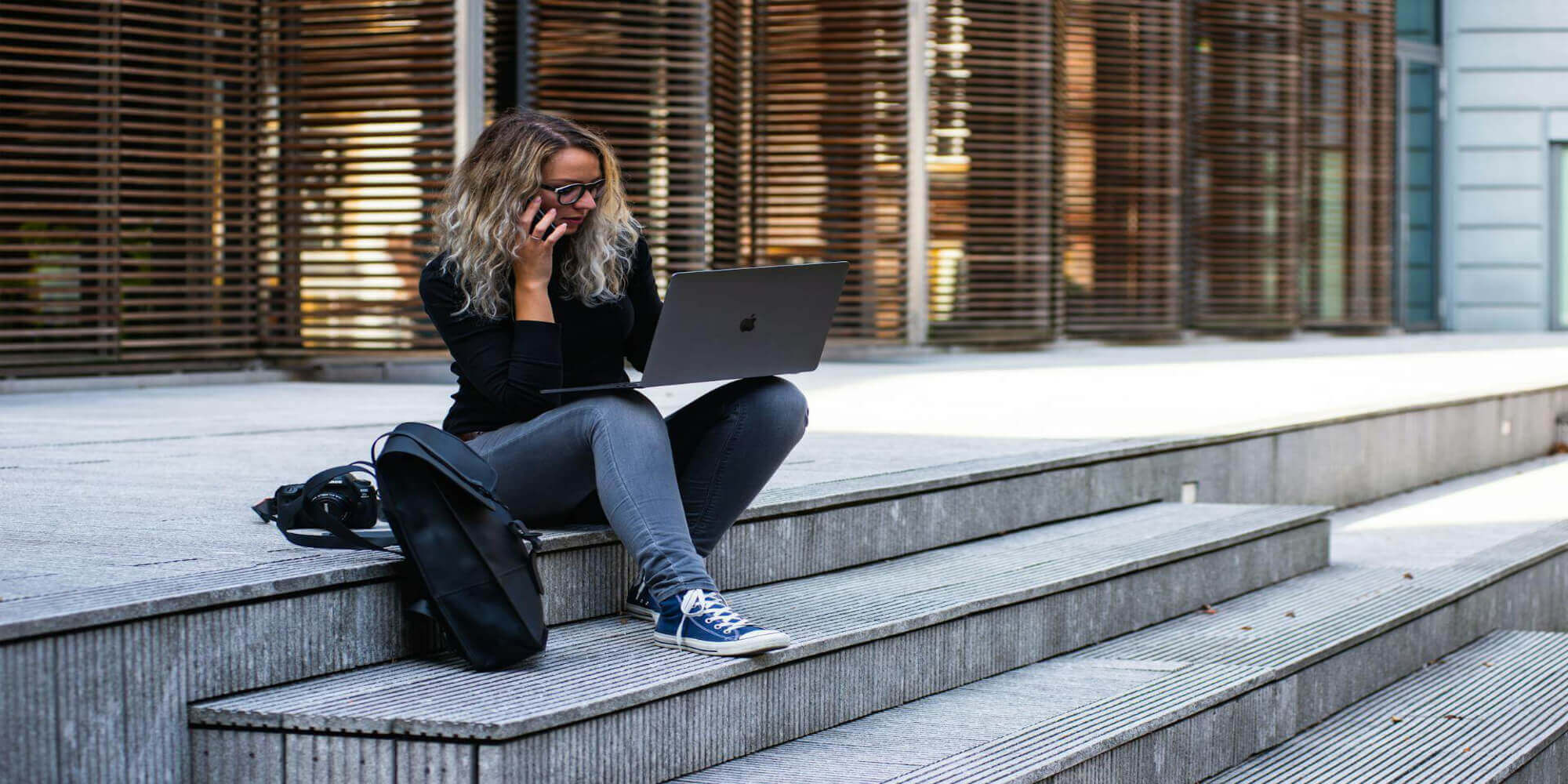 Eine Frau mit langen Haaren sitzt draußen auf einer modernen Treppe. Sie hält einen Laptop in der einen Hand und telefoniert mit einem Smartphone in der anderen Hand. Im Hintergrund ist ein modernes Gebäude zu sehen.